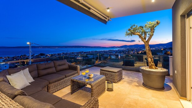 Ein Balkon mit einer Sitzecke, Tisch und Topfpflanze mit Blick auf eine Stadtlandschaft und das Meer in der Abenddämmerung.