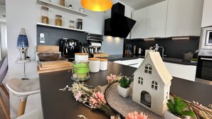 Een moderne keuken met witte kasten, een zwarte kookplaat en een koffieapparaat. Op het aanrecht staat een kleine kandelaar in de vorm van een huisje en een vaas met bloemen.