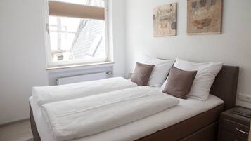 Ein ordentlich bezogenes Bett mit weißen Leinen und zwei Kissen, ein Nachttisch mit Fernbedienung und ein Fenster mit Jalousien.