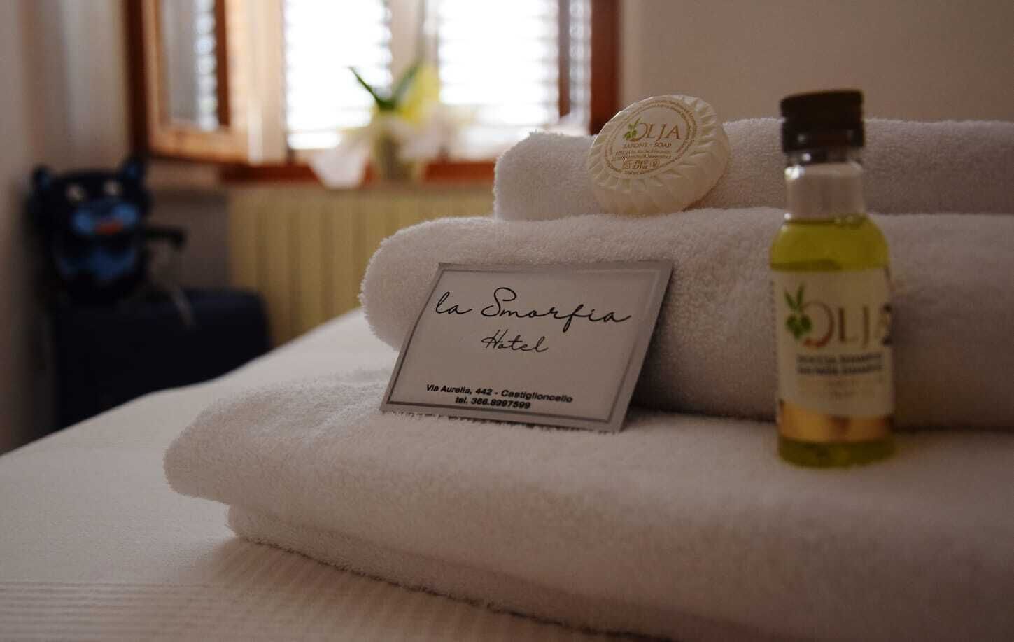 Una toalla blanca doblada con cuidado, una etiqueta que dice “la Smorfia Hotel” y una botella de aceite de oliva sobre una cama.