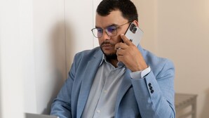 A person in a light blue suit sitting at a white desk with a laptop and a smartphone.