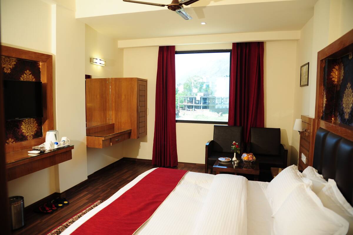 A hotel room with a large bed, a desk with a lamp, a chair, a TV, and a window with curtains.