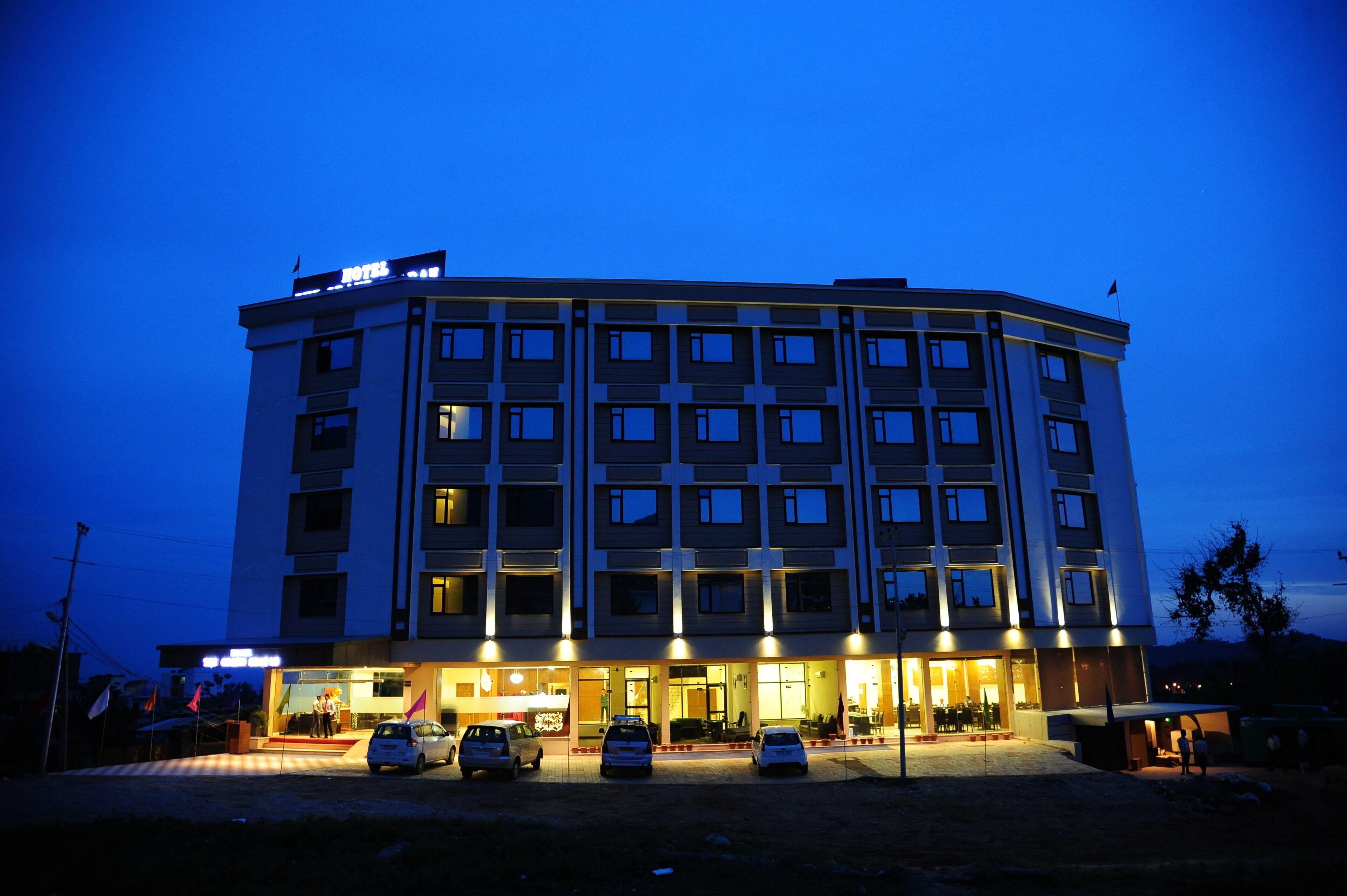 A multi-story hotel with a flat facade, multiple balconies, and a parking area with several cars.