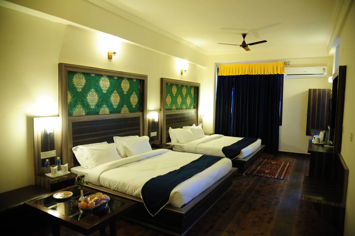 A hotel room with two beds, a ceiling fan, an air conditioning unit, and a nightstand with a lamp and a small table with a bowl of fruit.