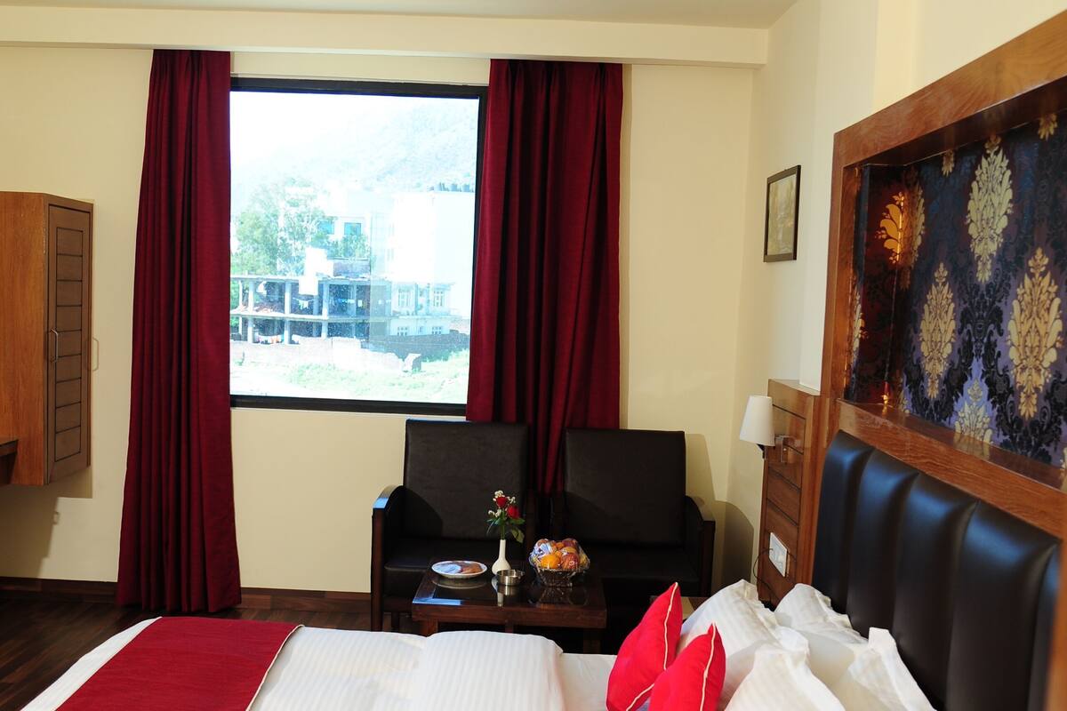 A hotel room with a bed, a desk, a chair, a window with curtains, a ceiling fan, and a view of the outside.