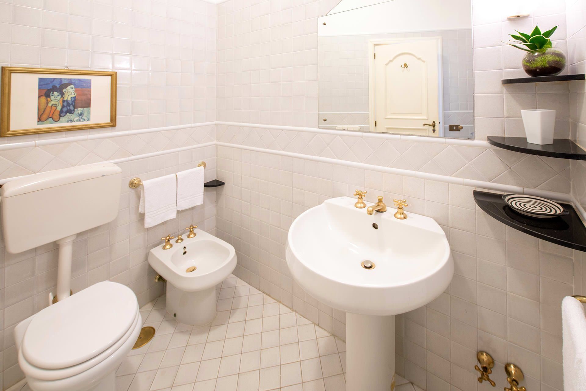 A bathroom with a toilet, bidet, and sink, all in white. There is a framed picture on the wall and a small plant on a shelf.