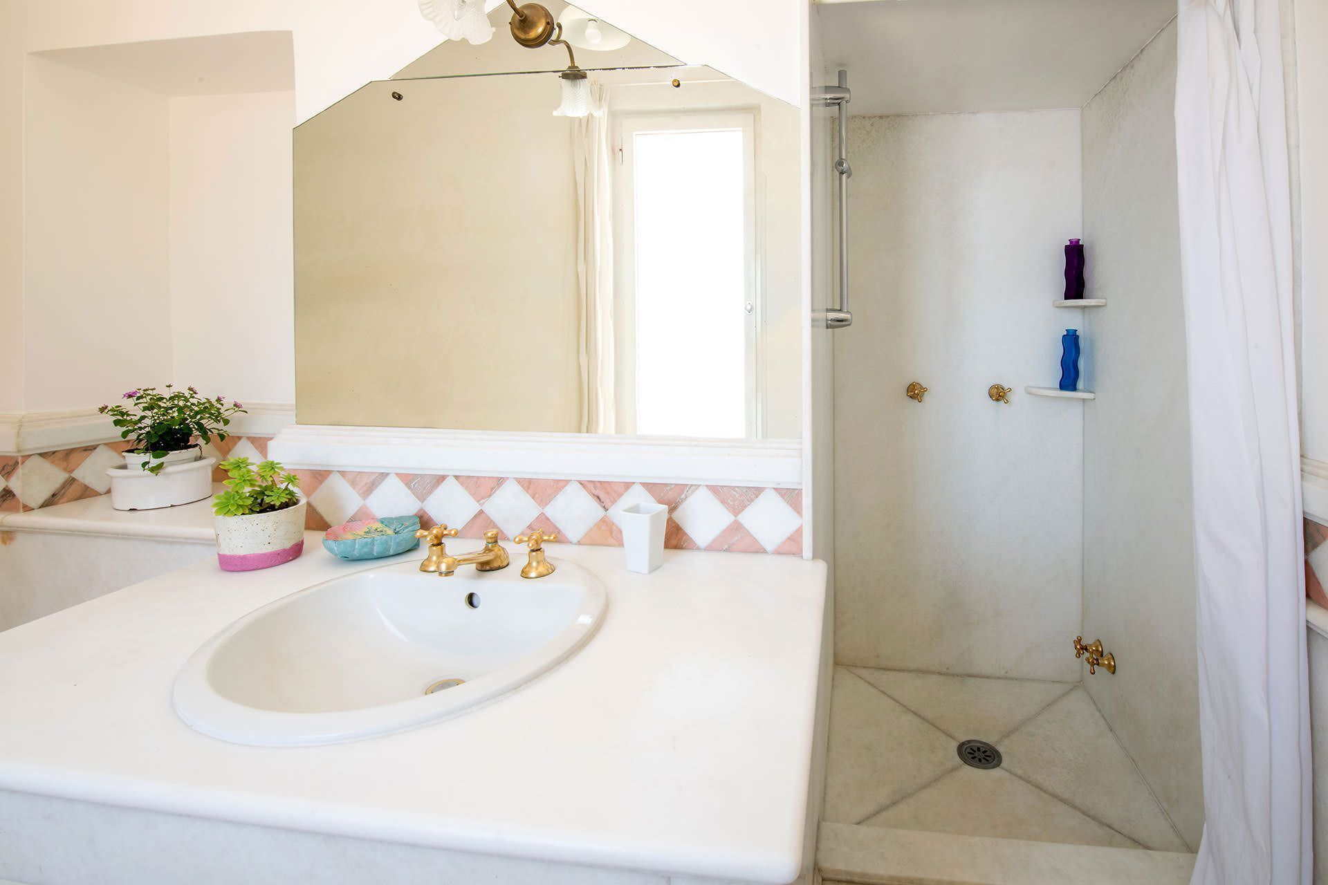 A bathroom with a white sink, two gold faucets, a mirror, and a shower area with a drain.