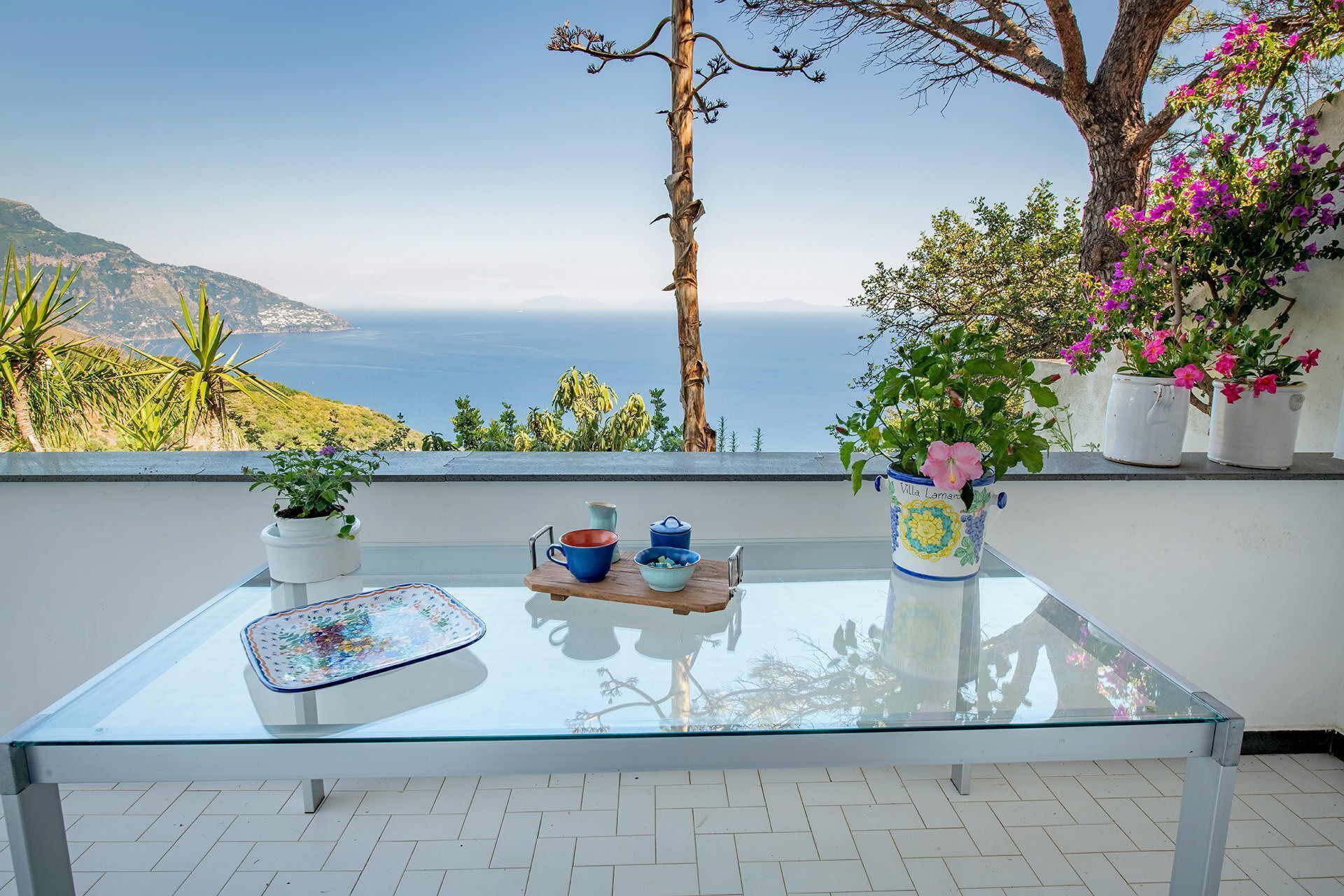 Uma mesa com tampo de vidro, toalha branca, duas plantas em vasos e uma tigela azul numa varanda com vista panorâmica.