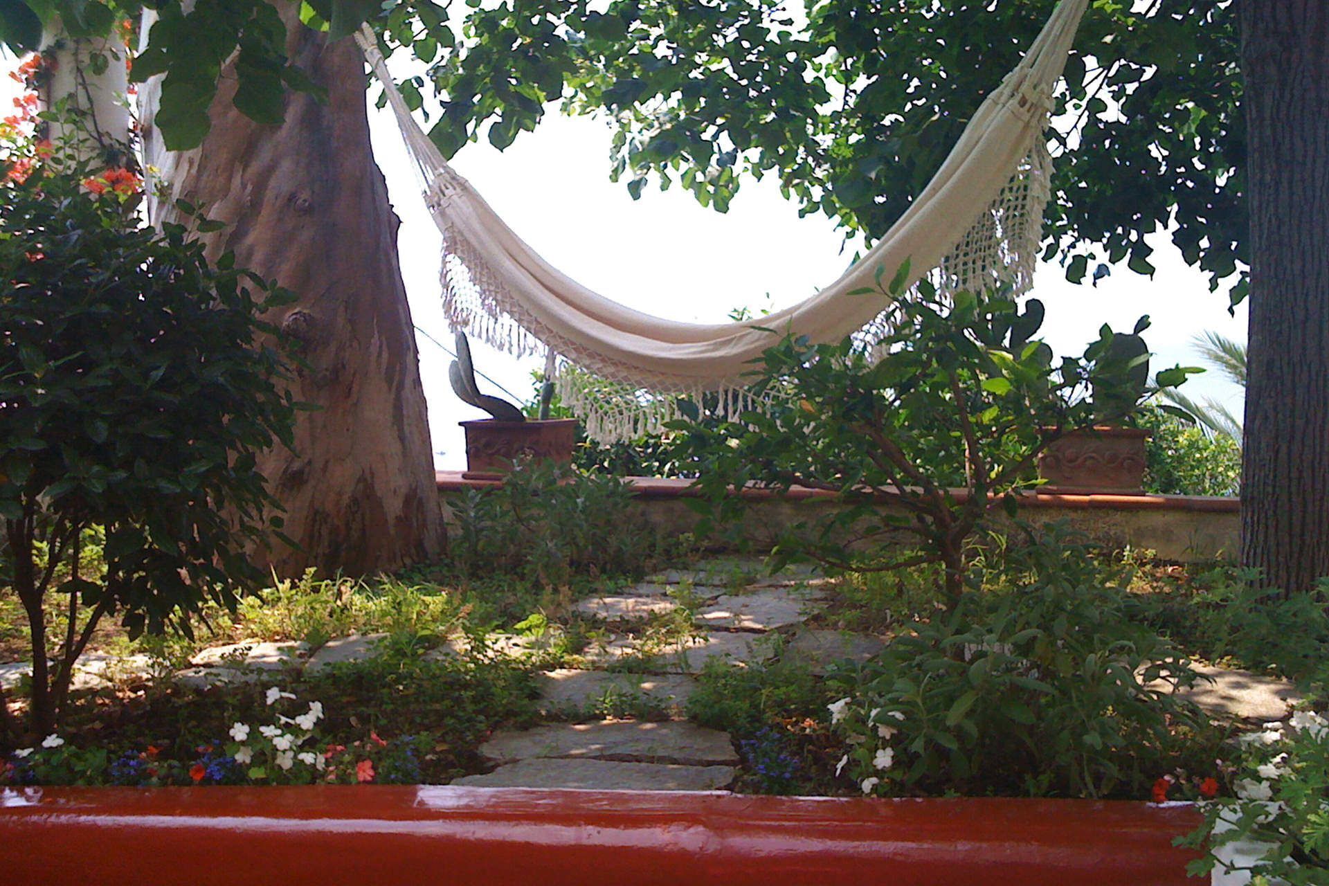Eine Hängematte, die zwischen Bäumen in einem Garten mit Steintreppen und Topfpflanzen hängt.