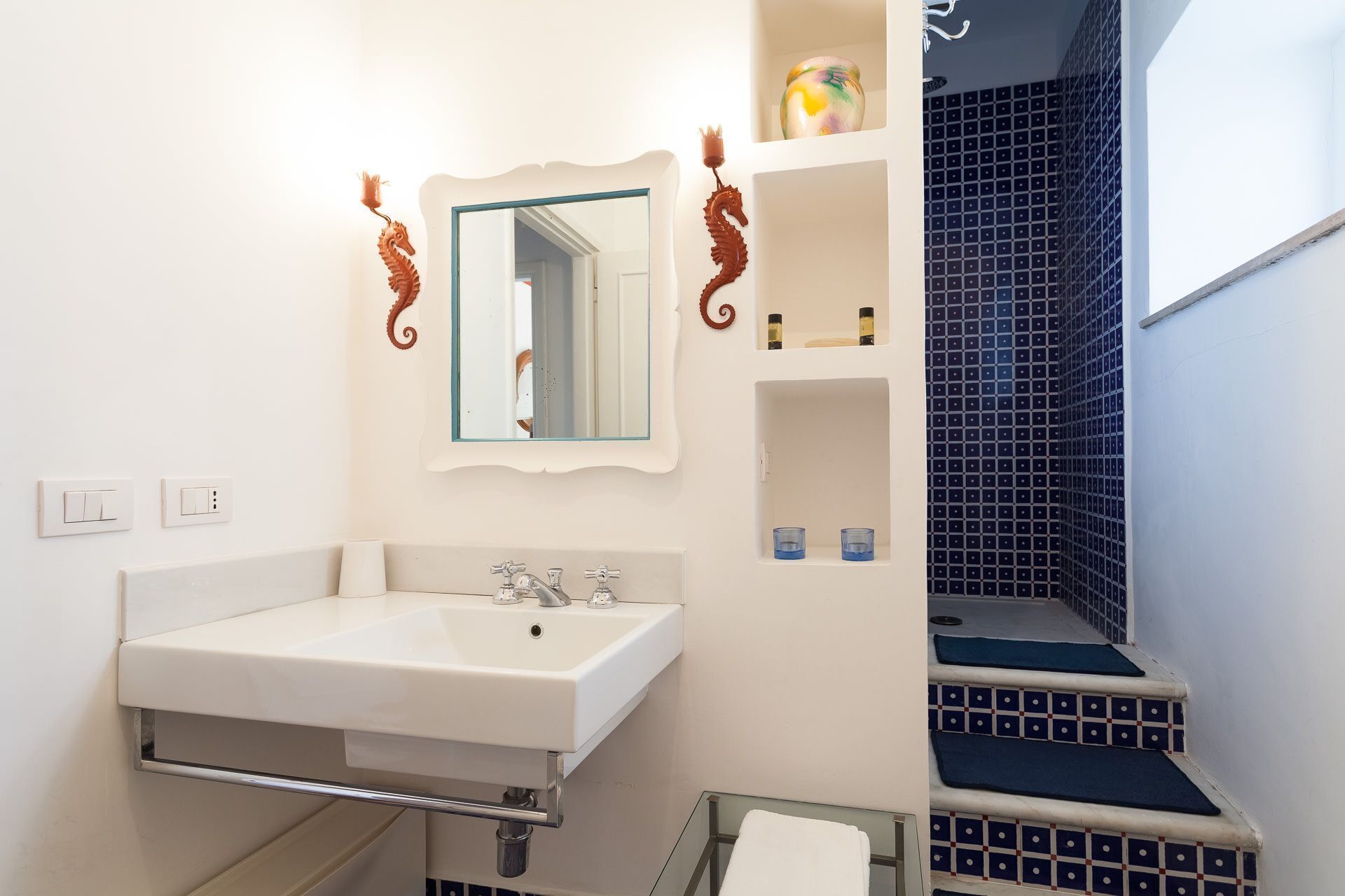 Une salle de bain avec un lavabo blanc, un miroir et des toilettes. Il y a deux étagères au-dessus du lavabo, l’une contenant des articles de toilette et l’autre un objet décoratif. Les murs sont blancs, et une partie du mur de droite est recouverte de carreaux bleus.