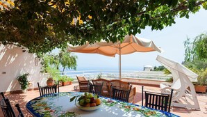 A round table with a fruit bowl, surrounded by chairs, under a large umbrella, with a view of the sea in the background. - Casa Dorothea 2 (Positano)