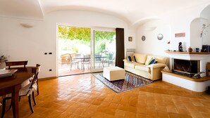 A living room with a sofa, a dining table with chairs, a fireplace, and a view through a glass door to an outdoor area. - Casa Dorothea 3 (Positano)