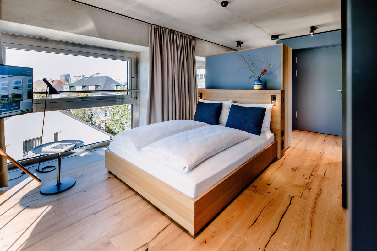 Ein modernes Hotelzimmer mit einem großen Bett, einem kleinen Schreibtisch mit einem Laptop, Blick auf die Stadt durch große Fenster und einem Balkon.