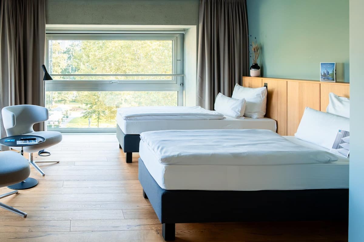Ein Hotelzimmer mit zwei Betten, einem Sessel, einem kleinen Tisch und einem Fenster mit Vorhängen.