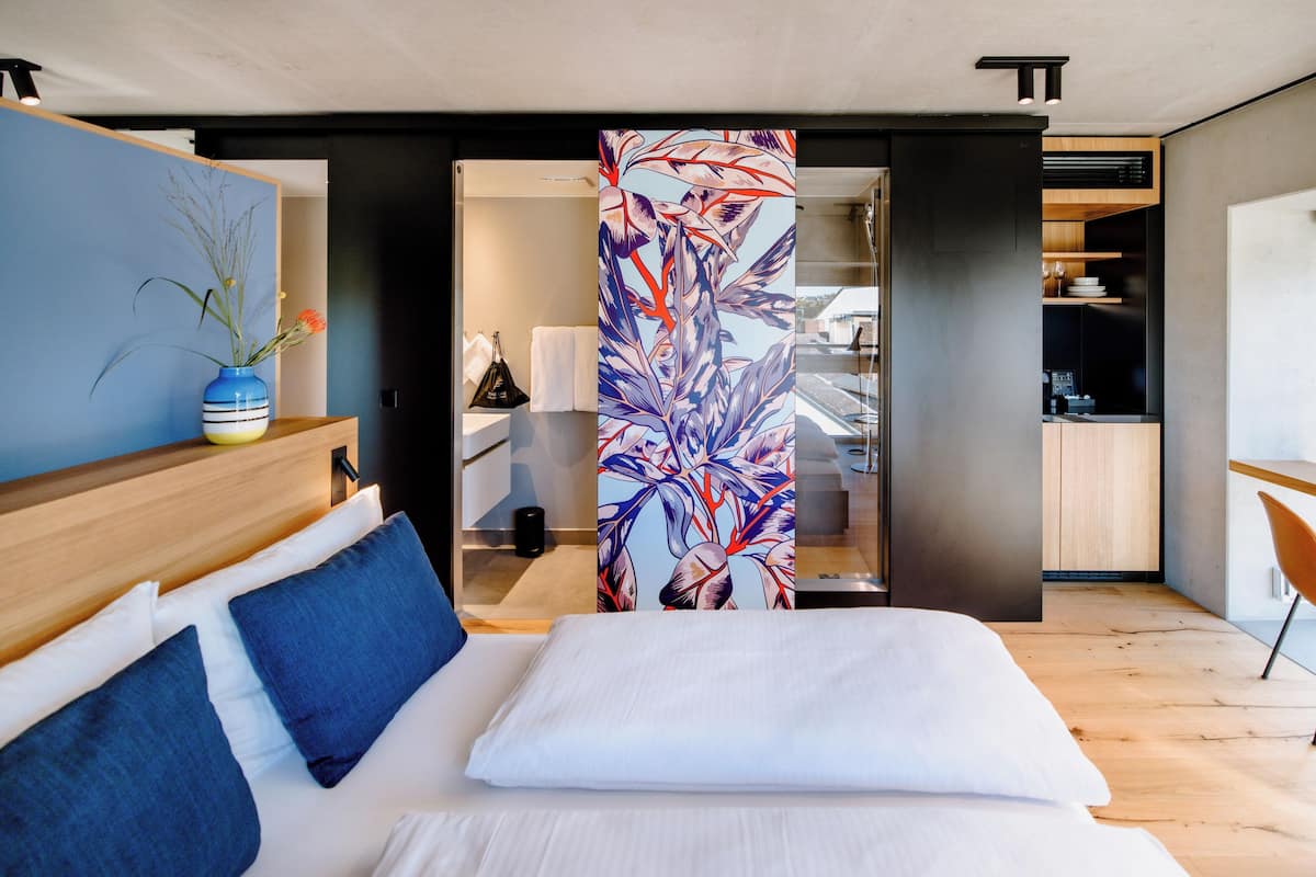 Ein modernes Hotelzimmer mit einem Bett, einer blauen Vase mit Blumen, einem hölzernen Kopfteil und Blick auf die Küche durch eine offene Tür.