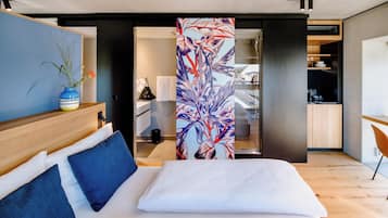 A modern hotel room with a bed, a blue vase with flowers, a wooden headboard, and a view of the kitchen through an open door.