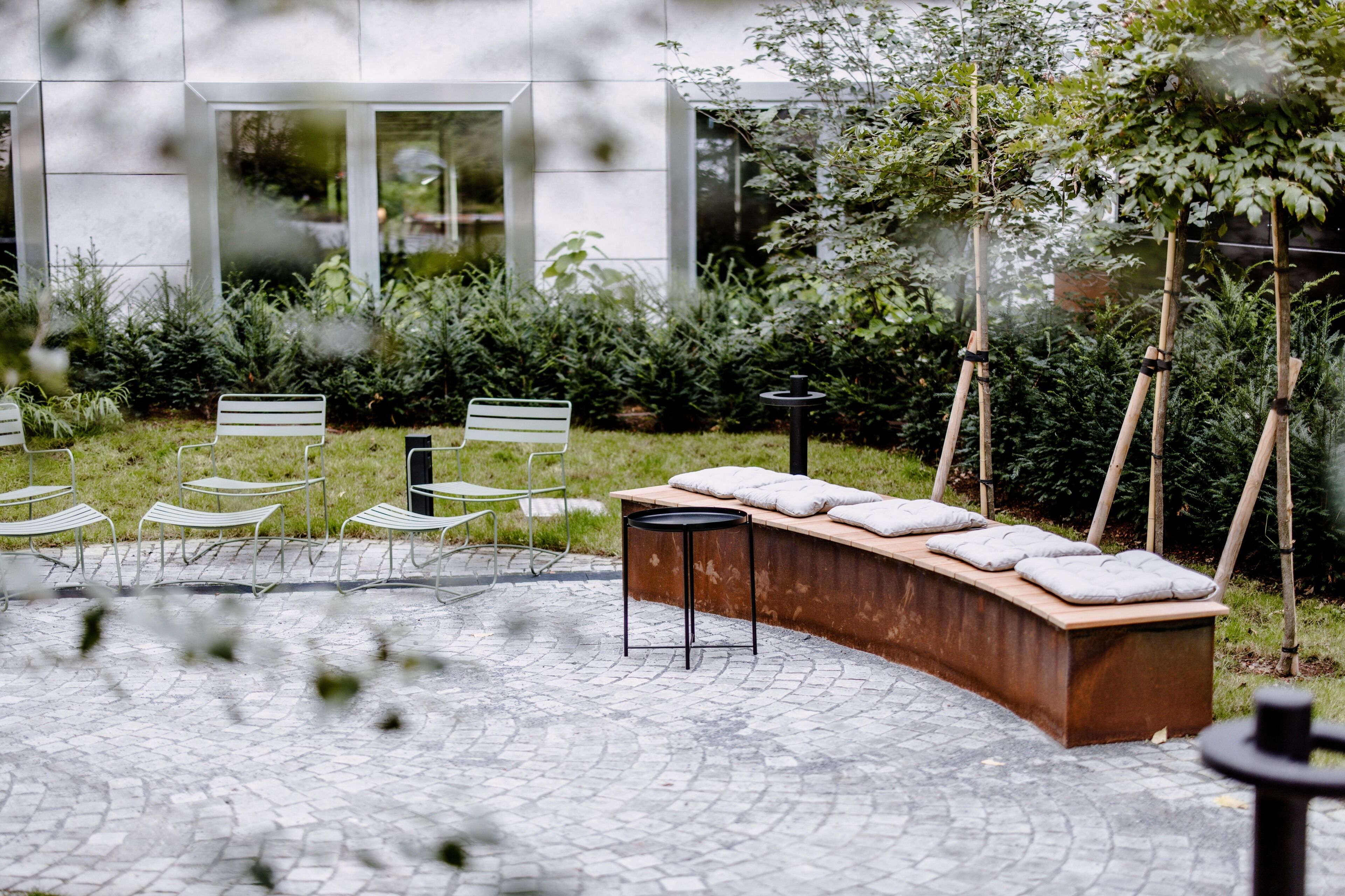 Une cour avec un espace aménagé comprenant un banc et des chaises, entourée de verdure et bordée d’un bâtiment doté de grandes fenêtres.