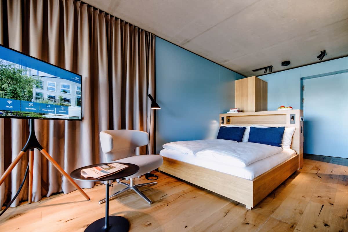 Ein modernes Hotelzimmer mit einem Holzbett, einem Schreibtisch, einem Stuhl und einem Fernseher, der eine Hotelwebseite zeigt.