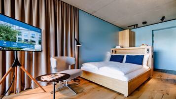 A modern hotel room with a wooden bed, a desk, a chair, and a TV displaying a hotel website.