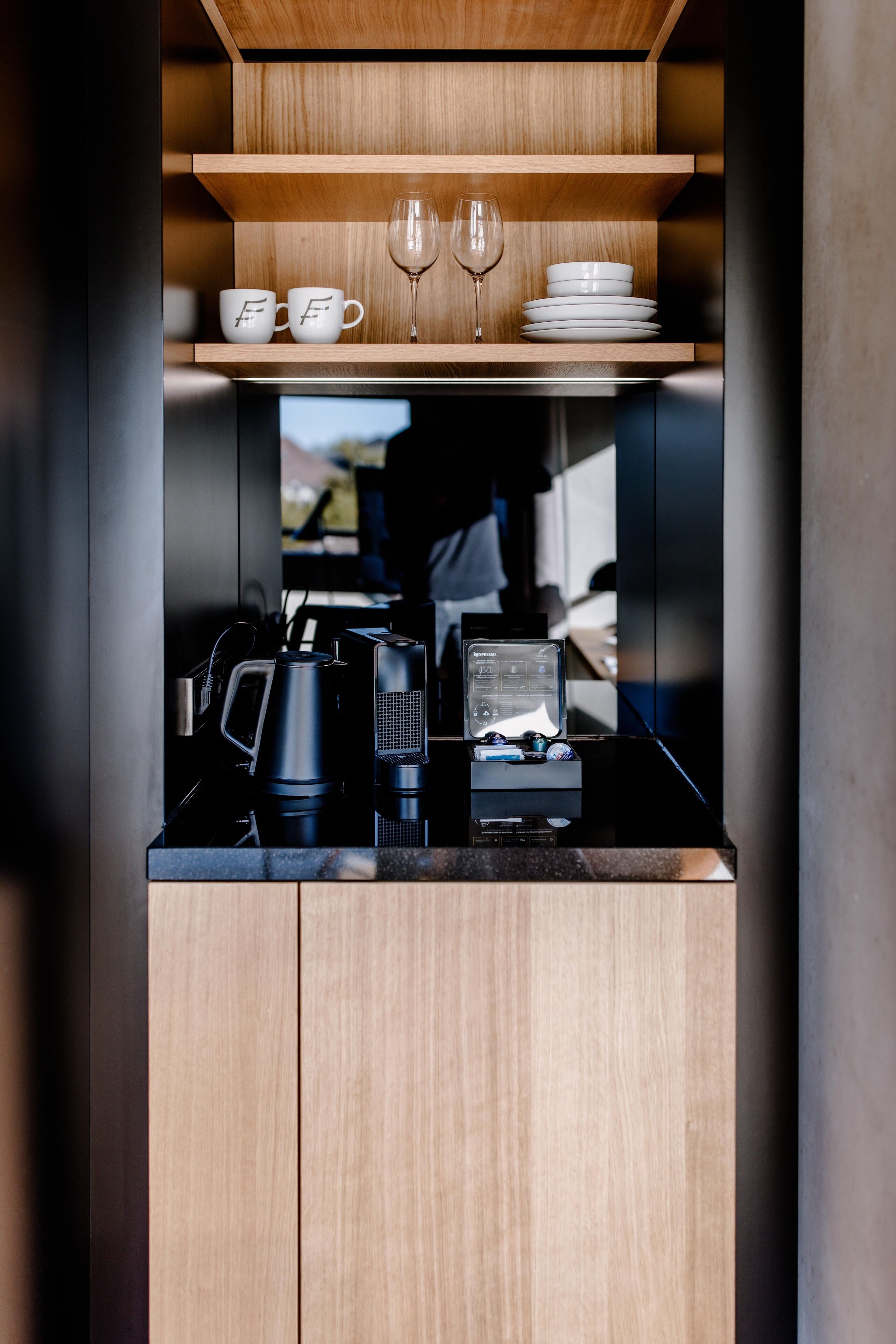 A compact kitchen with a wooden shelf holding cups and a wine glass, a coffee maker, and a small refrigerator.