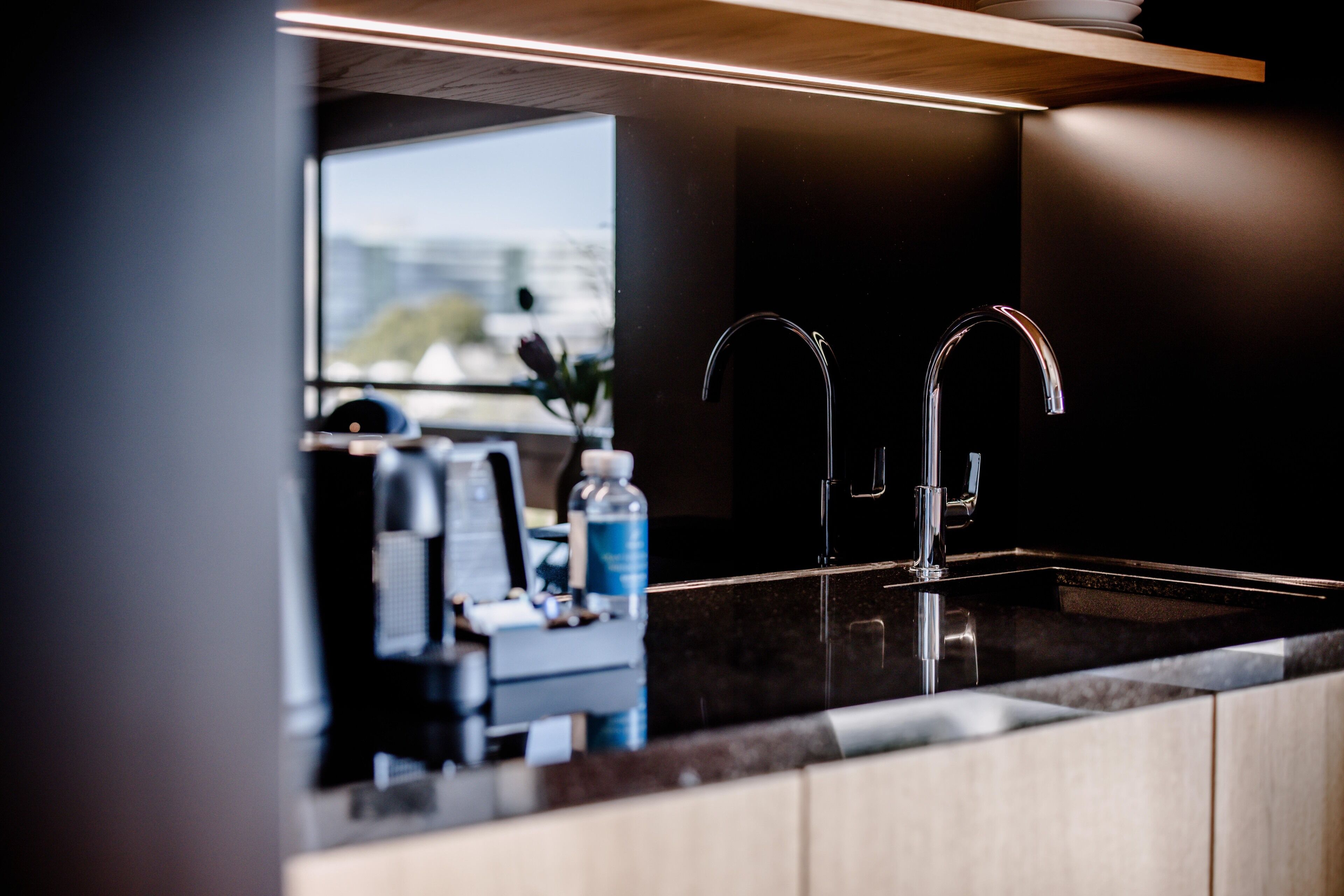 Una cocina moderna con encimera oscura, dos grifos de acero inoxidable y vistas al exterior a través de una ventana.