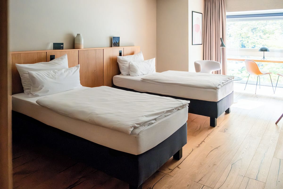 Ein Hotelzimmer mit zwei Betten, einem Holzkopfteil, einem kleinen Tisch mit Stuhl und einem Fenster mit Blick auf Grünflächen.