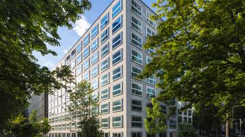 A modern multi-story building with a symmetrical facade, large windows, and surrounded by lush greenery.
