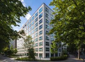 A modern multi-story building with a symmetrical facade, large windows, and surrounded by lush greenery.
