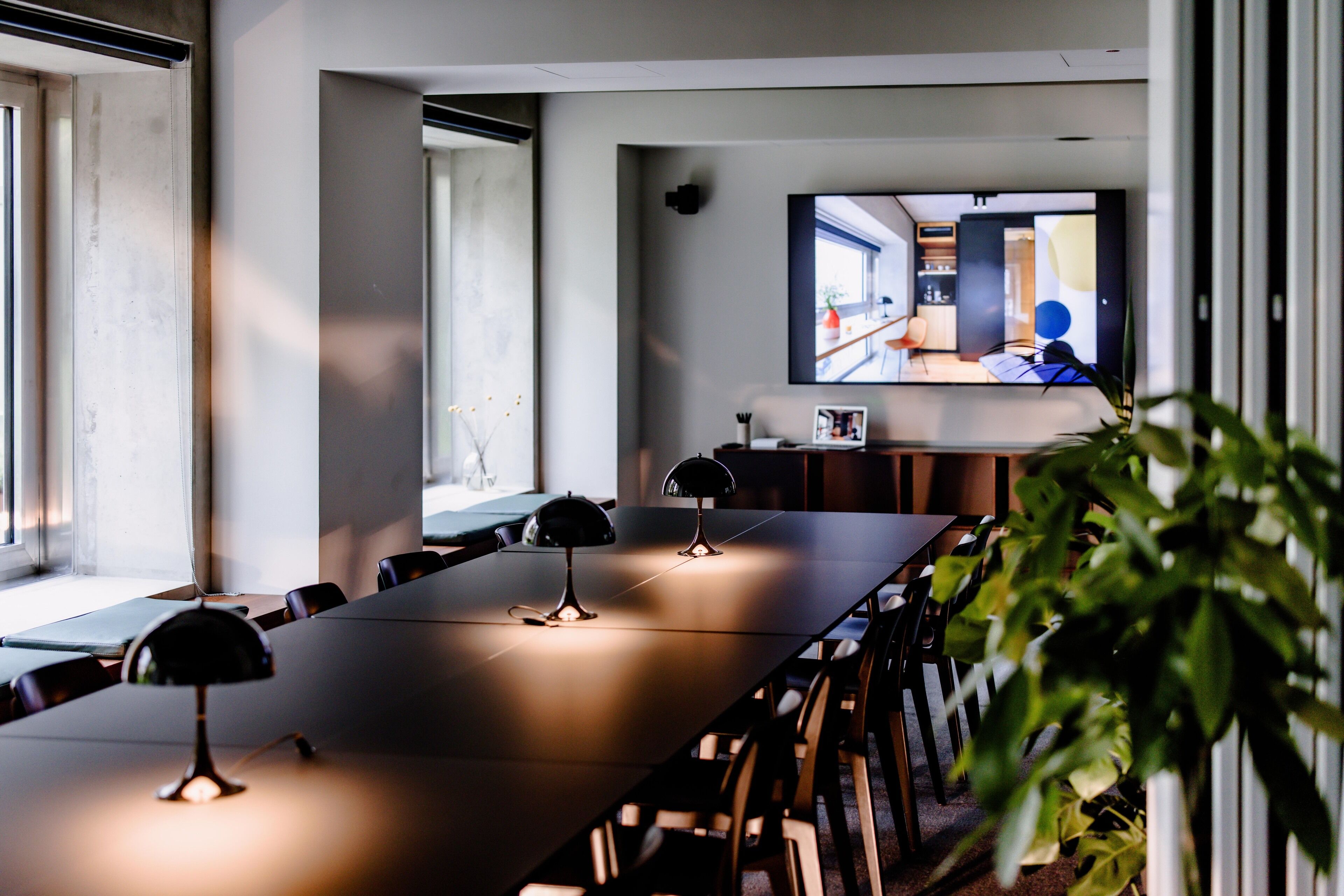 A conference room with a long table, chairs, and a large screen displaying a room.