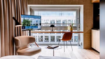 A modern room with a large window, a flat-screen TV, a wooden desk, a chair, and a small table with a book.