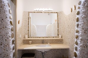 A bathroom with a speckled stone wall, a large mirror with lights, a sink, and a small cup on the counter. - Marvi House 1 (Mykonos)