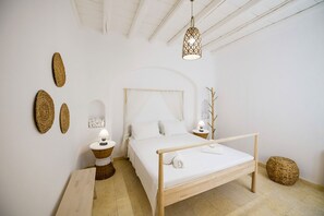 A minimalist bedroom with a wooden bed, a bedside table, a lamp, and woven wall decorations. - Marvi House 1 (Mykonos)
