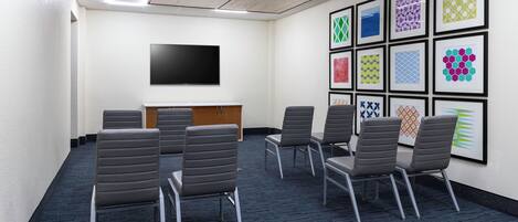 A conference room with a flat-screen TV, a wooden panel, and a row of grey chairs.