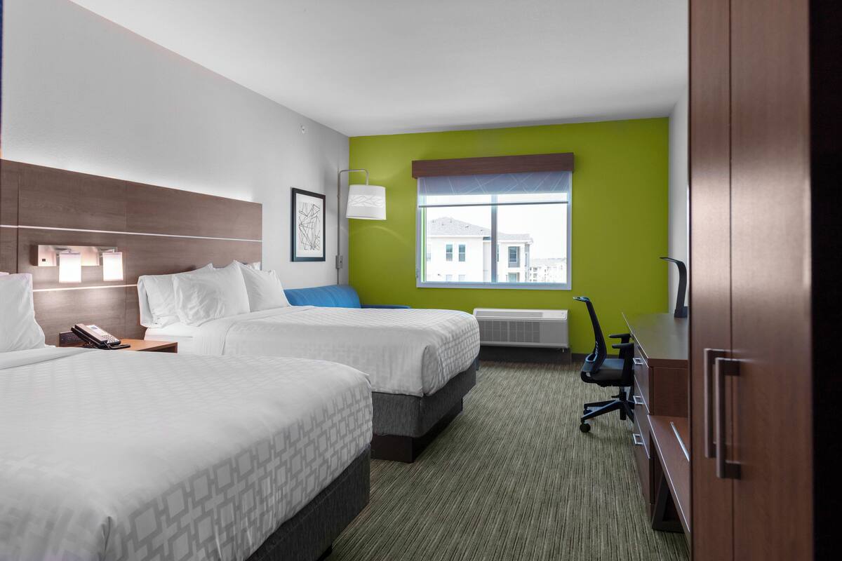 A hotel room with two beds, a desk, a chair, and a window with a view of buildings.