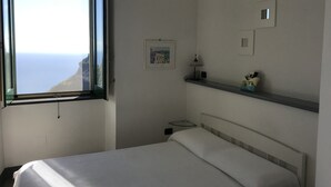 Un dormitorio con cama, mesita de noche, lámpara y estante con un objeto decorativo. Una ventana ofrece vistas al mar.