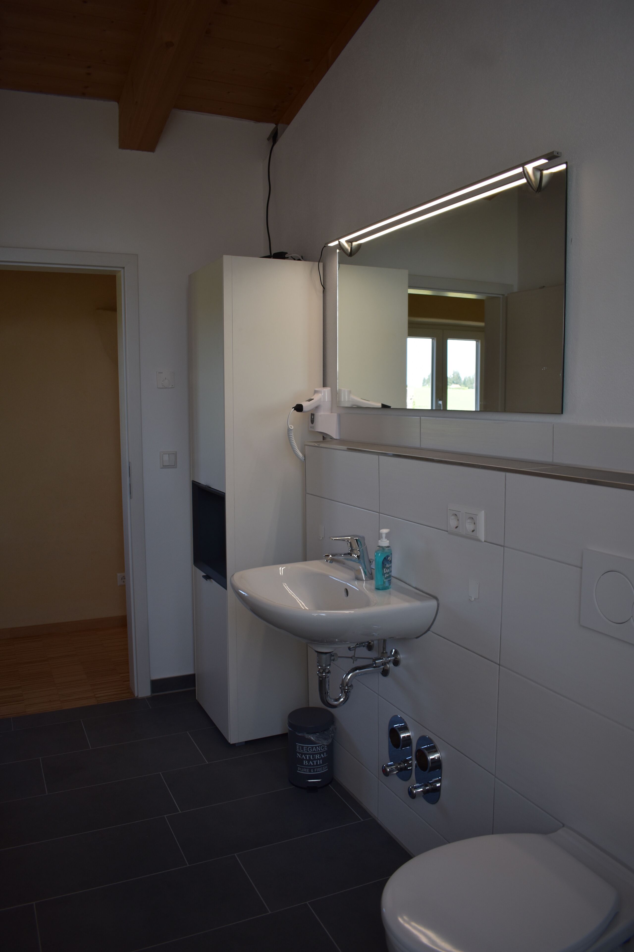A bathroom with a white sink, mirror, and toilet. There is a trash can and a bottle of hand soap on the sink.