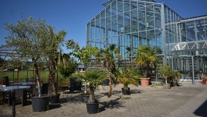 Een kas met glazen constructie, omringd door potplanten en buiten zitplaatsen.
