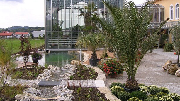 Een oranjerie met een glazen dak, omgeven door een tuin met diverse planten en een klein vijvertje.