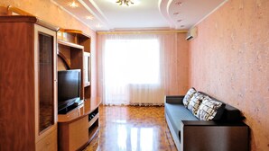 A room with a wooden entertainment unit, a flat-screen TV, a sofa with patterned cushions, a large window with sheer curtains, and a ceiling with recessed lighting. - Description of Staroreznichnaya 28 (Odessa)