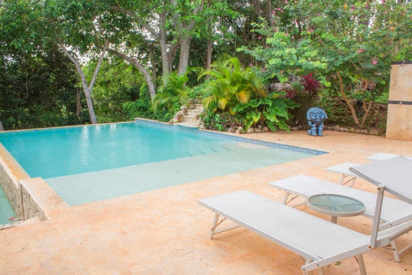 A rectangular swimming pool with a tiled deck, surrounded by lush greenery and a statue.