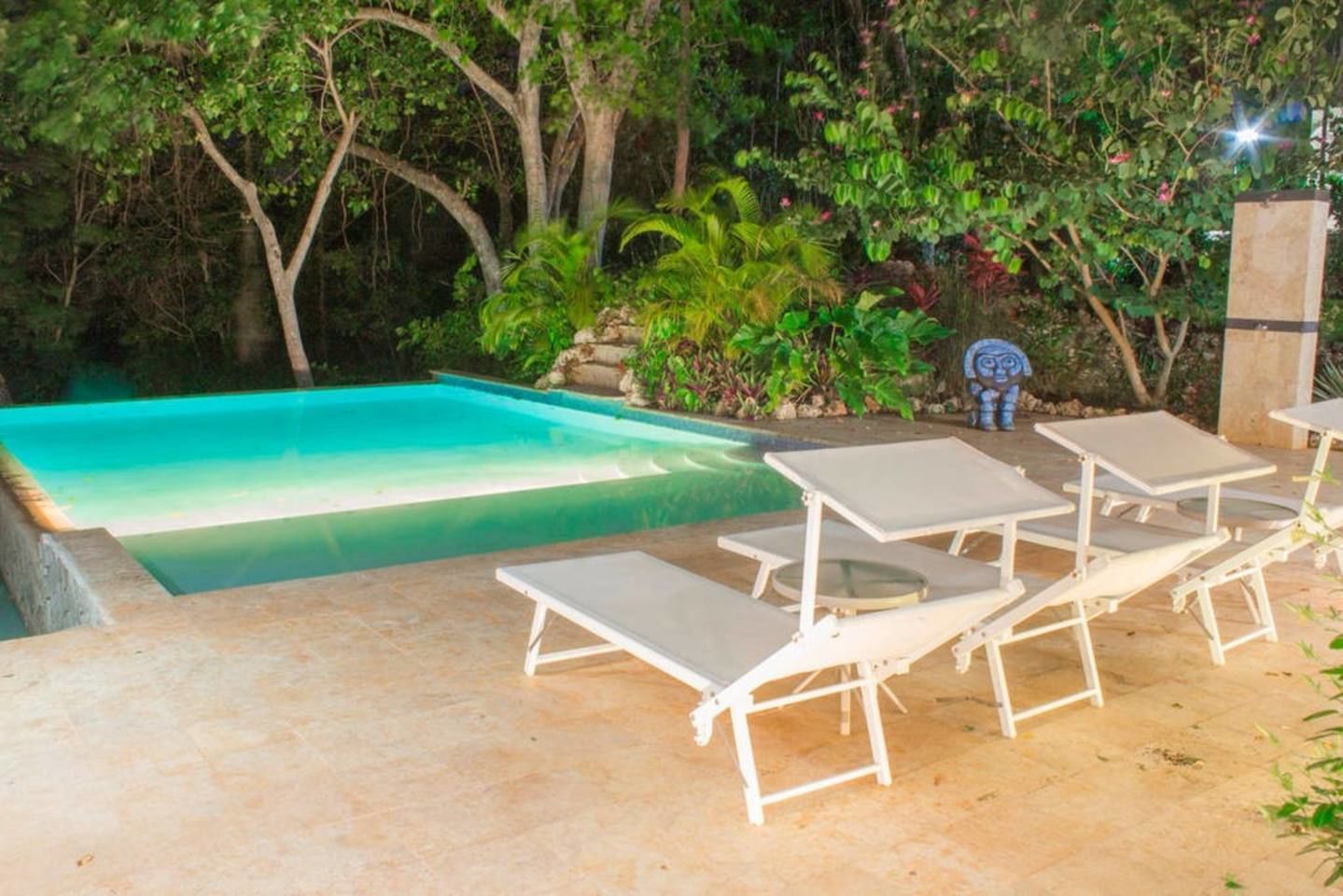 A swimming pool with a tiled deck, surrounded by lush greenery and a stone pillar.