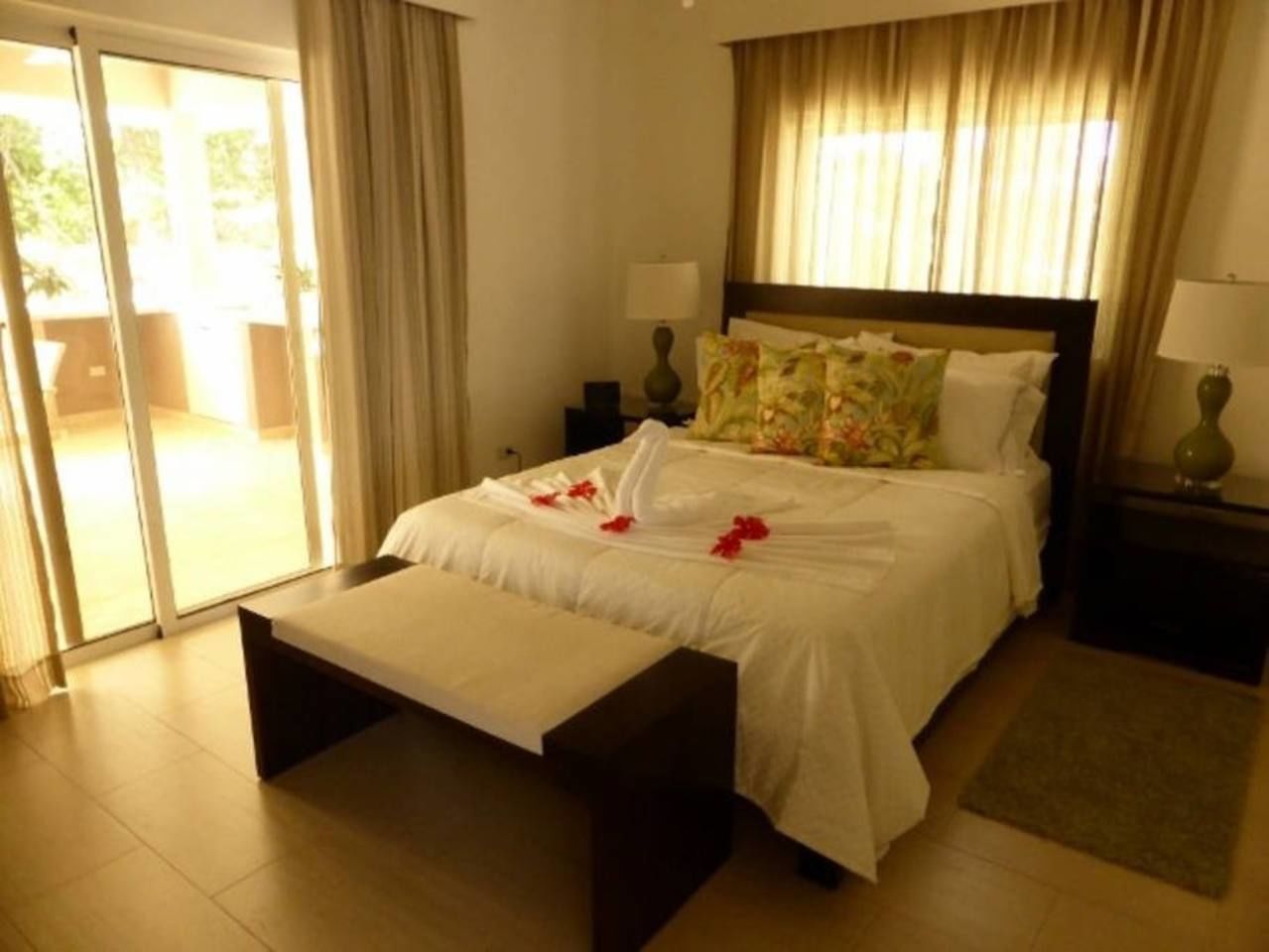 Habitación de hotel con cama, banco, mesita de noche, lámpara y vista al exterior a través de una puerta corredera.