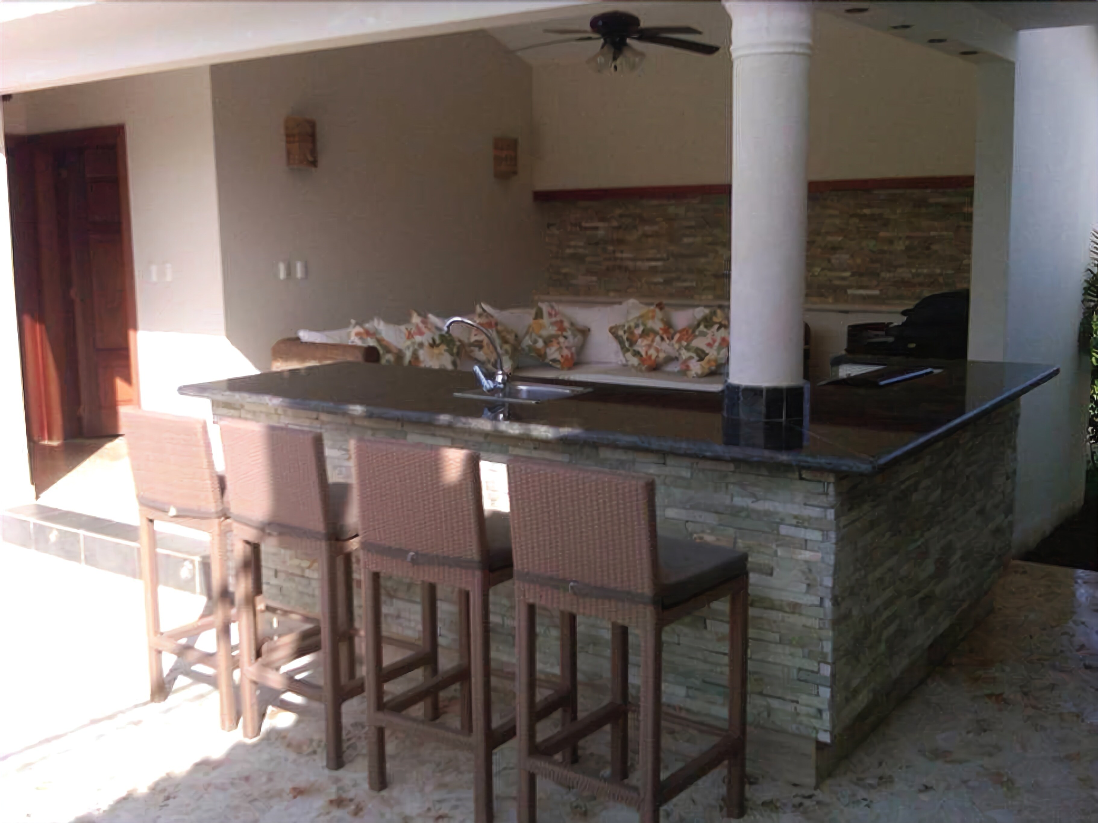 An outdoor kitchen area with a bar stool seating, a ceiling fan, and a stone backsplash.