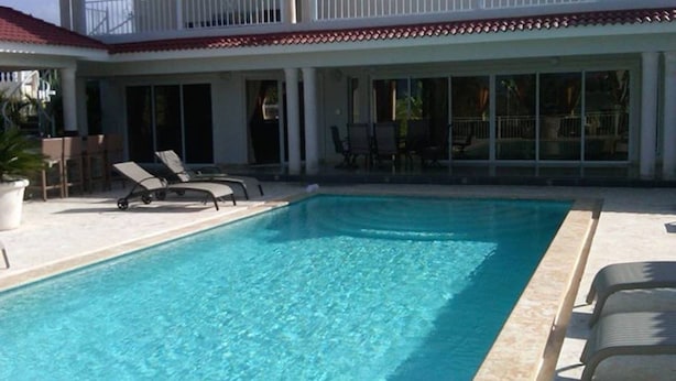 Una piscina rectangular con agua azul cristalina, rodeada de sillones de descanso y un porche techado.