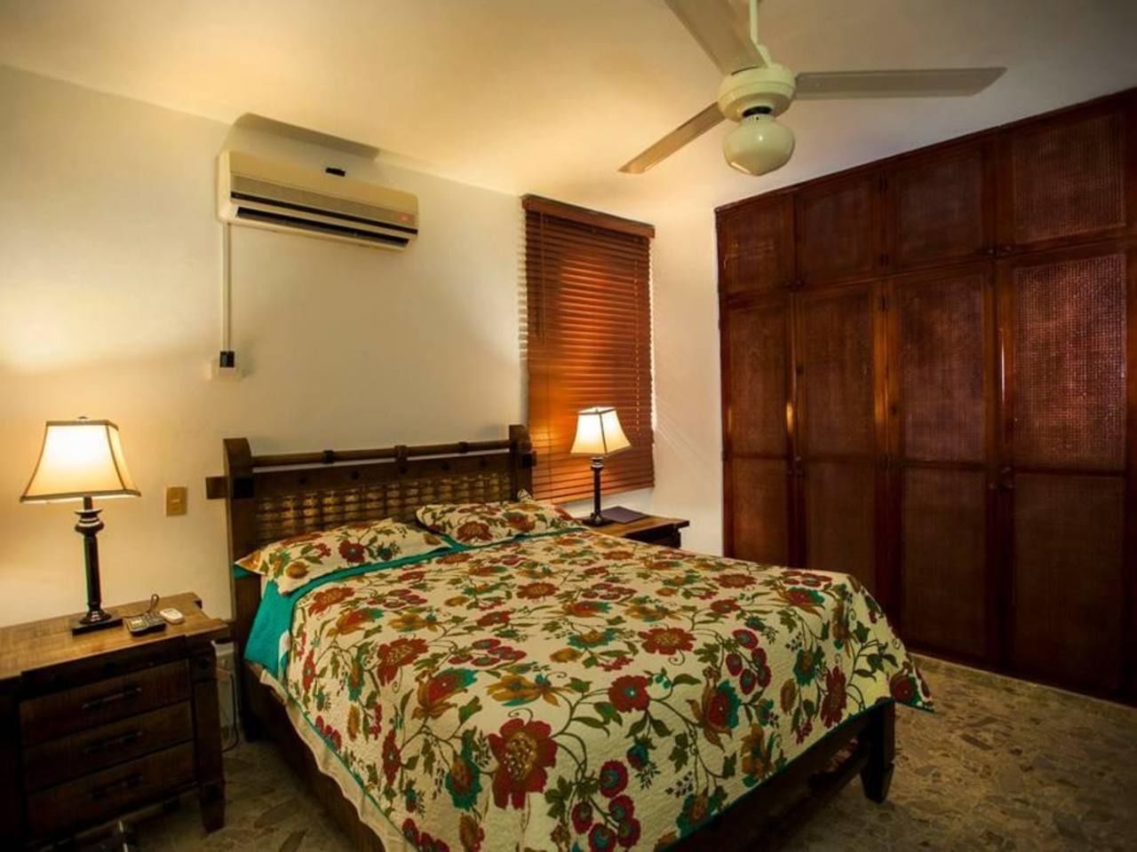 Quarto com cama, duas luminárias de cabeceira, ar condicionado, ventoinha no teto e guarda-roupa de madeira.