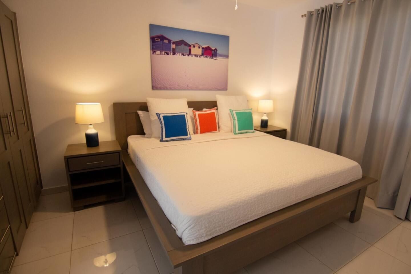 Una cama bien hecha con edredón blanco, dos mesitas de noche con lámparas, un cuadro de casas en la pared y una ventana con cortinas.