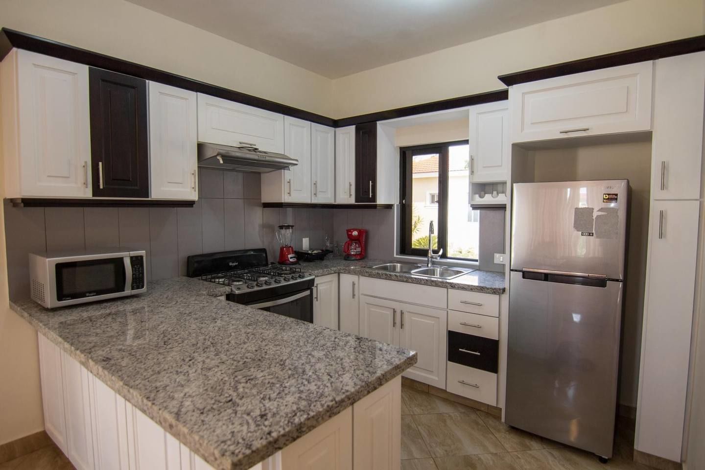 A kitchen with white cabinets, a granite countertop, a stainless steel refrigerator, a microwave, a stove, and a window with a view of the outside.