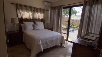 A bedroom with a bed, a nightstand, a lamp, a desk with a computer, and a sliding glass door leading to an outdoor area.