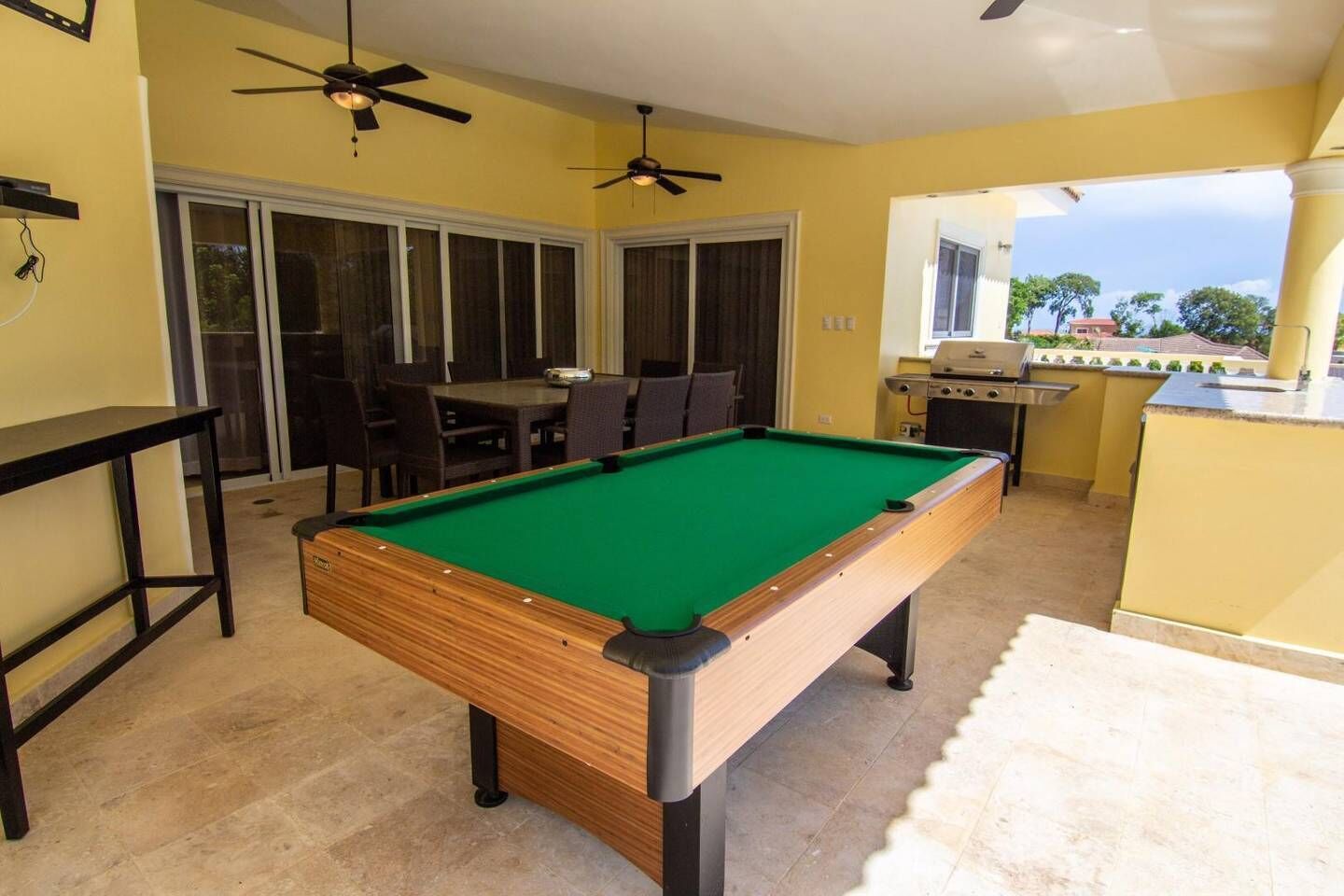 A pool table in a room with yellow walls, a dining area with chairs, and a kitchen area with a grill.