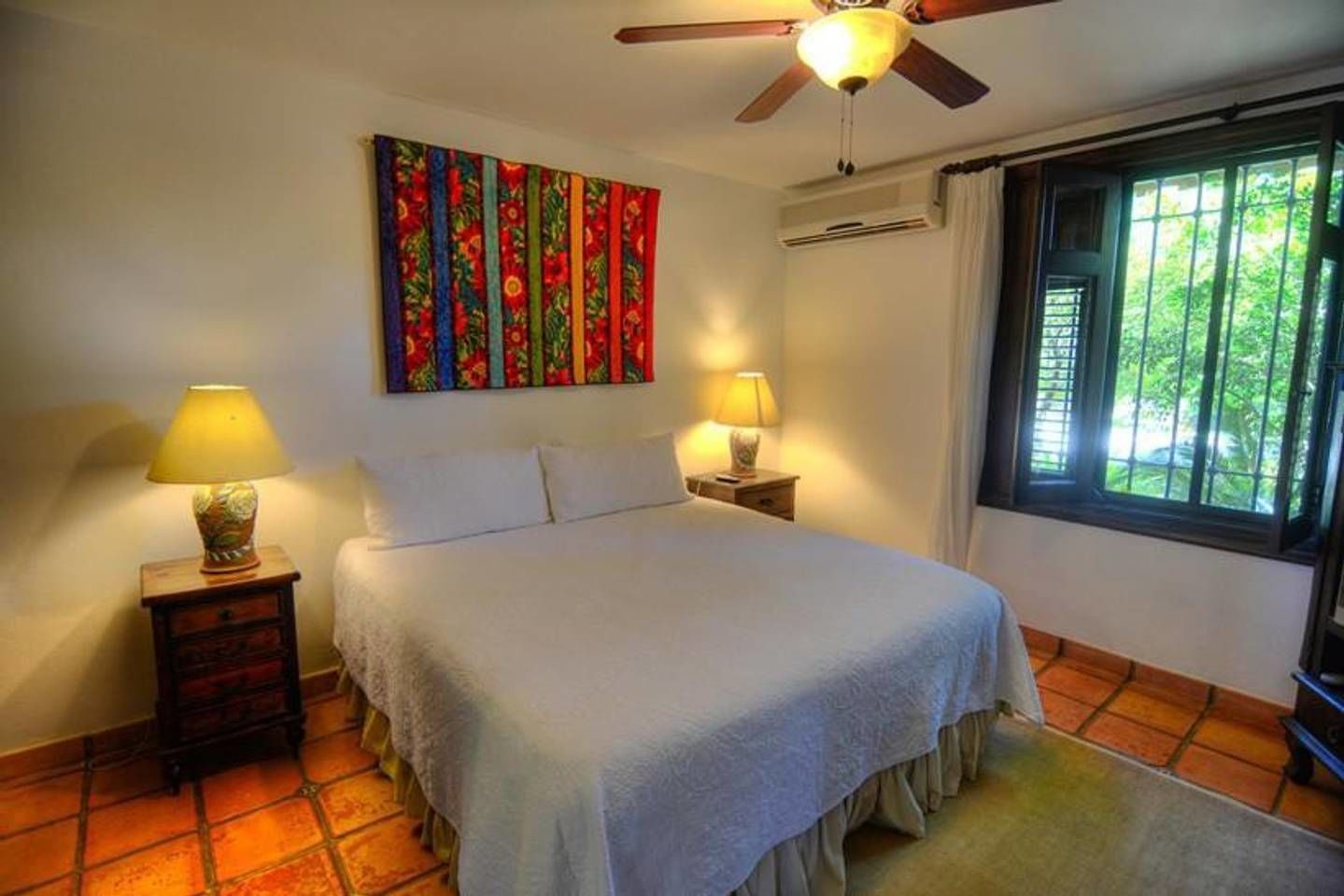 Un dormitorio con cama, dos mesitas de noche con lámparas, un ventilador de techo, una ventana con cortinas y un tapiz colorido en la pared.